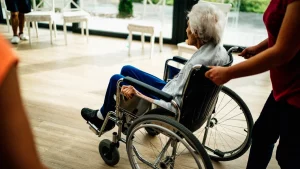 Pflegerin schiebt eine ältere Frau im Rollstuhl durch einen hellen Aufenthaltsraum
