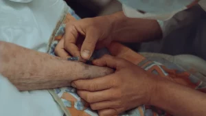 Angehörige und Pflegekraft halten die Hand eines pflegebedürftigen Menschen im häuslichen Pflegebett