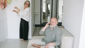 Seniorin mit Notrufknopf am Handgelenk in ihrer Wohnung, im Hintergrund die Basisstation des Hausnotrufs